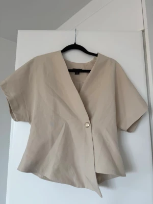 Beige kortärmad blus med knapp - Stilren beige blus med korta ärmar och v-ringning. Blusen har en asymmetrisk knäppning framtill med en dekorativ knapp. Tillverkad i ett lätt och mjukt tyg som ger en avslappnad men trendig look.