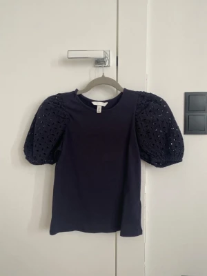 Svart topp med puffärmar från H&M - Trendig svart topp från H&M med puffiga, broderade kortärmar i spets och rund halsringning. Kroppen är i mjuk bomull och ärmarna har ett snyggt hålmönster som ger en cool touch till outfiten.