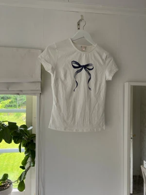 Vit t-shirt med blå rosett - Säljer en söt vit t-shirt med en blå rosett framtill. T-shirten har korta ärmar och rund halsringning. Perfekt för dig som gillar enkla men ändå unika detaljer. Materialet känns mjukt och skönt mot huden. Har några fläckar, se bilder.