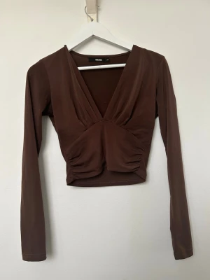 Brun Croppad topp med V-ringning - Snygg brun croppad topp från Bik Bok med djup v-ringning och långa ärmar. Toppen har rynkade detaljer framtill. Använd men i mycket fint skick. Storlek XS. 