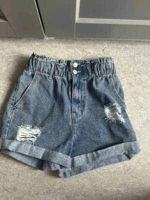 Blå jeansshorts - Säljer ett par blå denimshorts med hög midja och resår i midjan. Shortsen har slitna detaljer framtill, uppvikta benslut och klassiska fem fickor. Perfekta för sommaren och ger en avslappnad vibe.