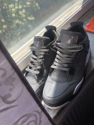 Air Jordan 4 Retro grå/svart - Snygga Air Jordan 4 Retro sneakers i grått och svart med vita och gula detaljer. Skorna har klassisk meshpanel, robust gummisula och ikonisk Jumpman-logga på plös och sula. Perfekta för dig som gillar streetwear och vill sticka ut med en tidlös siluett.