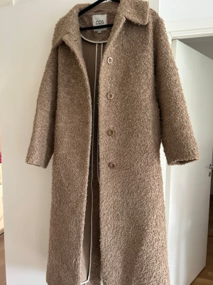 Beige teddyjacka från COS - Lång, beige teddyjacka från COS med klassisk krage och knäppning framtill. Jackan har ett mysigt, fluffigt material och ett matchande knytbälte i midjan för en snygg siluett. Perfekt för kalla dagar när du vill vara både varm och trendig.