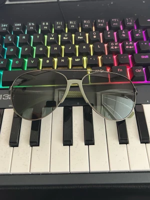Emporio Armani aviator solglasögon - Snygga aviator solglasögon från Emporio Armani med silvrig metallbåge och mörktonade linser. Skalmarna har en grön detalj med textur som ger en unik touch. Perfekta för dig som vill ha en klassisk men modern look.