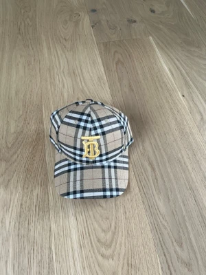 Burberry beige rutig keps - Säljer en klassisk Burberry keps i beige med det ikoniska rutmönstret och ett gult broderat 'B' framtill. Kepsen har böjd skärm och justerbar rem bak. Tillverkad i bomull och perfekt för dig som vill ha en tidlös accessoar med lyxig känsla.