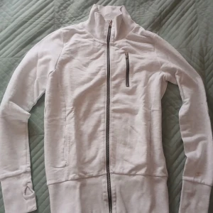 Vit zip hoodie med hög krage - Säljer en tight, ljusgrå hoodie med hel dragkedja, hög krage och en bröstficka med dragkedja. Tröjan har långa ärmar med tumhål och ribbade muddar. Den är lite mindre än storlek M, men materialet är mjukt och perfekt för chilla dagar eller träning.