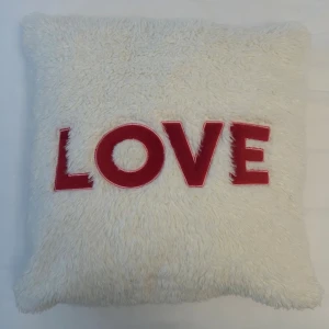 Vit fluffig kudde med LOVE-tryck - Mysig fyrkantig kudde i vitt fluffigt material med texten LOVE i stora röda bokstäver på framsidan. Kudden har en mjuk och gosig känsla och passar perfekt som inredningsdetalj. Kudden har måtten 45x45 cm. 