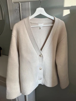 Beige kofta i ullmix med knappar - Snygg beige kofta i ullmix med djup V-ringning och stora pärlemorknappar framtill. Koftan har breda muddar och en avslappnad passform, perfekt att slänga över en t-shirt eller topp. Enkel och stilren design som passar till det mesta. Nypris 1500kr