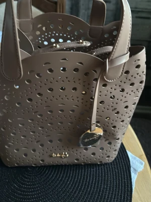 Beige handväska från Michael Kors - Snygg beige handväska från Michael Kors i konstläder med coolt laserutskuret mönster och guldfärgade detaljer. Väskan har två handtag, magnetknäppning och en rund MK-tag i metall. Perfekt för dig som gillar trendiga accessoarer.