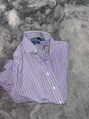 Polo Ralph Lauren Striped Shirt – Purple & Green - Klassisk och superfresh skjorta från Polo Ralph Lauren i en unik och iögonfallande färgkombination med lila, gröna och vita ränder. Modellen är "Regent Classic Fit" vilket ger en skön och avslappnad siluett. • Märke: Polo Ralph Lauren • Modell: Regent Classic Fit • Färg: Lila, Grön, Vit • Passform: Classic Fit. Sitter grymt! • Detaljer: Broderad logga på bröstet och button-down krage. • Skick: Toppskick, nytvättad och fräsch! 