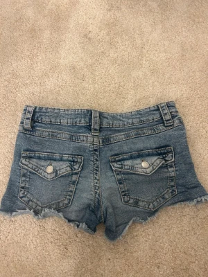 Blå jeansshorts med fransig kant - Säljer ett par blå jeansshorts med fransig kant och råa avslut. Shortsen har fickor både fram och bak, bälteshällor och knappstängning. Perfekta för varma dagar och en avslappnad stil.