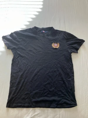 Svart t-shirt med broderad logga - Svart t-shirt från Vespucci med rund hals och korta ärmar. På bröstet finns en broderad logga i guld och rött med text och emblem. Tillverkad i mjuk bomull för en bekväm känsla. Perfekt till jeans eller shorts.