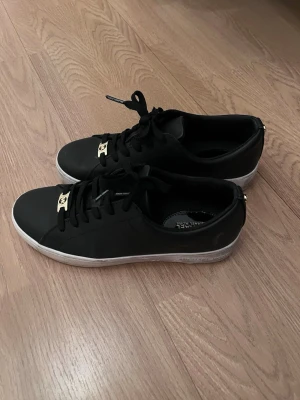 Svarta sneakers från Michael Kors - Stilrena svarta sneakers från Michael Kors med vita sulor och guldfärgade detaljer på snörningen. Skorna har en klassisk låg modell och är tillverkade i ett slätt material som ger en clean look. Perfekta för dig som gillar en enkel men lyxig stil.