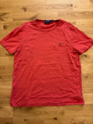 Röd t-shirt från Polo Ralph Lauren - Snygg röd t-shirt från Polo Ralph Lauren i modellen Custom Slim Fit. Klassisk rund halsringning och ikoniska broderade loggan på bröstet. Tillverkad i mjuk bomull som känns skön mot huden. Perfekt till jeans eller shorts för en lopp clean look.