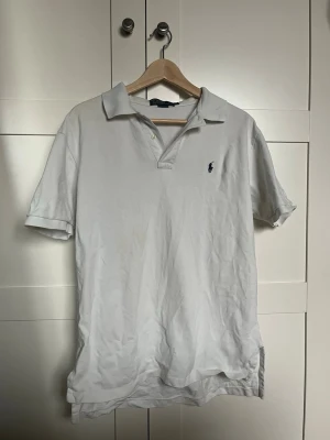 Vit pikétröja från Ralph Lauren - Klassisk vit pikétröja från Ralph Lauren med korta ärmar och krage. Tröjan har två knappar vid halsen och den ikoniska broderade loggan på bröstet. Tillverkad i mjuk bomull för en bekväm känsla. Den är lite uppriven på den vänstra sidan men ingenting man märker.