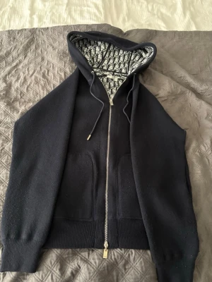 Marin hoodie med dragkedja  - Snygg marin blå hoodie med dragkedja och broderad logga på fickan. Huvan har ett unikt mönstrat foder och justerbara snören med metalldetaljer. Tillverkad i mjukt material och har ribbade muddar samt två fickor framtill.
