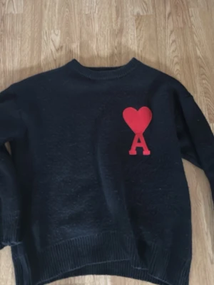 Svart stickad tröja Ami Paris - Säljer en svart stickad tröja från Ami Paris med röd hjärtlogga och bokstaven A på bröstet. Tröjan har rund halsringning, ribbade muddar och är gjord i mjukt material. Perfekt för dig som gillar stilrena plagg med en twist.