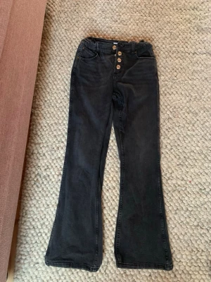 Svarta bootcut jeans med knappar - Säljer ett par svarta bootcut jeans med hög midja och fyra silverfärgade knappar i gylfen. Jeansen har klassiska fickor fram och bak samt bälteshällor. Materialet är stretchigt denim som sitter snyggt och bekvämt.