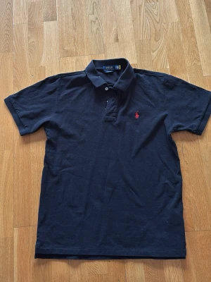 Svart pikétröja från Polo Ralph Lauren - Klassisk svart pikétröja från Polo Ralph Lauren med korta ärmar och röd broderad logga på bröstet. Tillverkad i mjuk bomull och har en normal passform med krage och knappar vid halsen. Perfekt till jeans eller shorts.