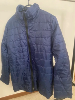 Mörkblå pufferjacka med dragkedja - Snygg mörkblå pufferjacka med hög krage och hel dragkedja framtill. Jackan har ett quiltat mönster och är tillverkad i ett glansigt syntetmaterial som håller värmen bra. Perfekt för kalla vinterdagar och enkel att matcha med olika outfits.