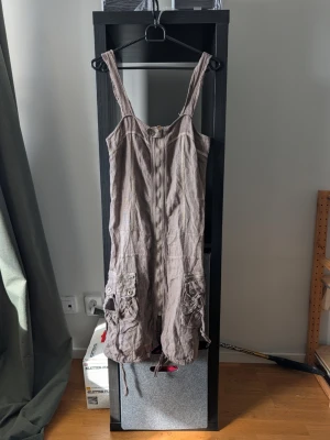 Beige linneklänning med fickor - Säljer en beige kortklänning i linne med breda axelband och dragkedja framtill. Klänningen har två stora fickor med knappar och snörning nedtill för en avslappnad look. Perfekt för varma dagar och enkel att styla. 100% lin