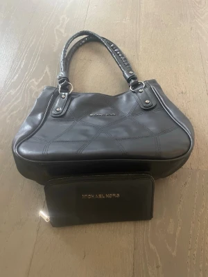 Svart handväska från Michael Kors - Snygg svart handväska från Michael Kors i glansigt läder med dubbla handtag och silverfärgade detaljer. Väskan har diskret logga framtill och stilrena sömmar som ger en cool look. Perfekt för dig som gillar klassisk men trendig design.