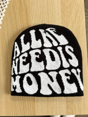 Svart mössa med vit text - Säljer en svart mössa med stor vit text i grafisk stil där det står 'ALL WE NEED IS MONEY'. One size. Unisex. Skickar gärna fler mössor i bundle. Mössan har en klassisk form och är tillverkad i mjukt stickat material, perfekt för dig som vill sticka ut med ett statement-plagg.