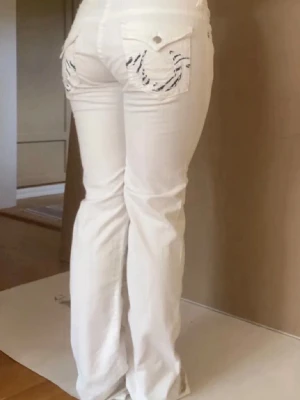 Lågmidjade vita jeans True Religion - Såå snygga vita lågmidjade bootcut jeans från True Religon. Har zebra mönster bak på fickorna!  Har används några enstaka gånger och är nästan som nya! Han en pytteliten fläck på vaden där bak som enkelt går att tvätta! ( Det är nog någon form av lera från utomhus när man har gått)  Storlek: 31, skulle säga att der motsvarar storlek 40 🥰 Midjemått: 40 cm