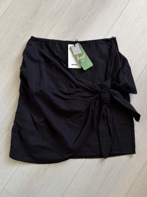 Svart omlottkjol från Bik Bok - Trendig svart omlottkjol från Bik Bok i luftig linnemix. Kjolen har en snygg knytning i sidan som ger en cool och avslappnad look. Perfekt till varma dagar och enkel att styla med olika toppar.