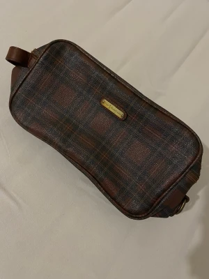 Hanväska från Polo Ralph Lauren - Säljer en snygg handväska från Polo Ralph Lauren i brunt och mörkgrönt rutigt mönster. Väskan är av konstläder med detaljer i brunt Perfekt för att samla småsaker eller använda som sminkväska.