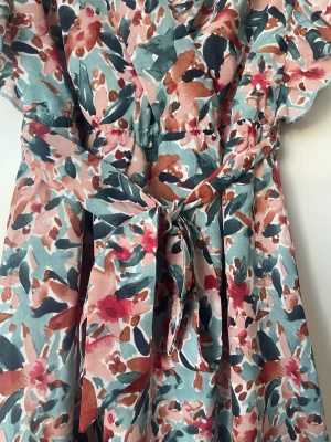 Blommig kortklänning med volang - Söt kortklänning med blommigt mönster i rosa, grön, vit och röd. Klänningen har v-ringning, volangdetaljer vid ärmar och nederkant samt ett knytband i midjan. Perfekt för sommarens alla tillfällen!