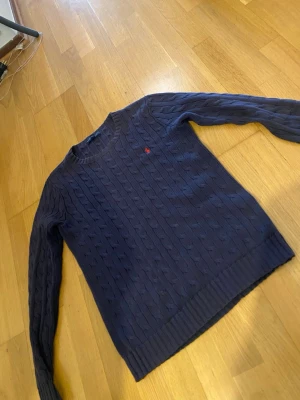 Mörkblå kabelstickad tröja Polo Ralph Lauren - Mörkblå kabelstickad tröja från Polo Ralph Lauren med rund halsringning och röd broderad logga på bröstet. Tröjan har långa ärmar och ribbade muddar vid ärmslut och nederkant. Klassisk och stilren design i mjukt material.