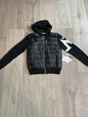Svart pufferjacka från Moncler - Snygg svart pufferjacka från Moncler med huva och dragkedja framtill. Jackan har ett quiltat parti på kroppen, släta ärmar och en stor vit blixtdetalj på ena ärmen. Moncler-logga på axeln och praktiska fickor med dragkedja. Passar även de som bär S 