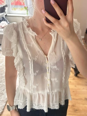 Vit spetsblus med volanger - Romantisk vit blus med broderade blommor, volangdetaljer och knappar framtill. Blusen har korta ärmar med spets och en lätt rynkad nederkant. Tillverkad i ett tunt, genomskinligt material som ger en luftig känsla.