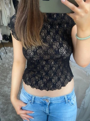 Svart spetsblus med kort ärm - Säljer en svart, genombruten spetsblus med korta ärmar och vågig kant nertill. Blusen är figurnära och har ett blommigt spetsmönster som ger en elegant vibe. Perfekt att styla med jeans eller kjol för en trendig look.