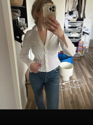 Vit figursydd skjorta med knappar - Säljer en vit, figursydd skjorta med v-ringning och knappar framtill. Skjortan har lång ärm och markerad midja som ger en snygg siluett. Perfekt att styla med jeans eller kjol för en clean och trendig look. Finns ute på Vinted med 