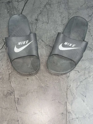 Gråa Nike badtofflor - Säljer ett par gråa Nike badtofflor med vit logga på ovandelen och Nike-tryck längs sulkanten. Tofflorna har öppen tå och är tillverkade i ett lätt syntetmaterial som torkar snabbt. Perfekta för stranden eller gymmet.