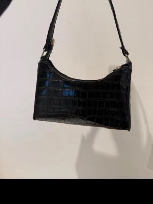 Svart axelväska i krokopräglat skinn - Snygg svart handväska med axelrem i krokopräglat konstläder. Väskan har en rektangulär form och stängs med dragkedja upptill. Perfekt accessoar för att lyfta din outfit med en trendig touch.