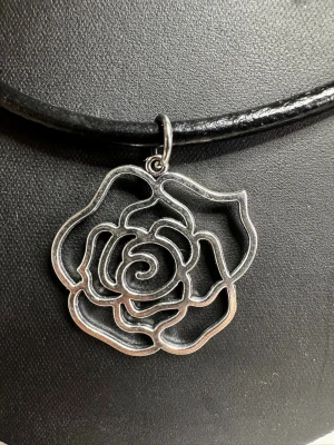 Halsband med rosformad silverberlock - Snyggt halsband med svart läderrem och en stor, rosformad berlock i silverfärgad metall. Berlocken har en öppen, stilren design som ger ett modernt intryck. Perfekt accessoar för dig som gillar unika smycken med blommotiv.