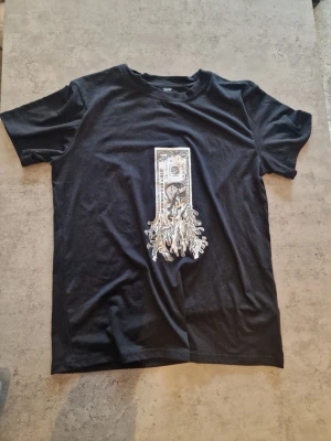 Svart t-shirt med dollartryck - Cool svart t-shirt med ett stort tryck av en brinnande dollarsedel på framsidan. T-shirten har klassisk passform och rund hals. Perfekt för dig som gillar streetwear och vill sticka ut med en unik design. Står small i storlek men upplever att den är lite stor själv säga att den passar medium