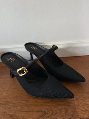 Svarta pumps med gulddetalj från Cosy Island  - Nya! Aldrig använda. Strl us 8 motsvarar strl 38. Stilrena svarta pumps från Cosy Island med spetsig tå och elegant rem över foten. Skorna har en snygg guldfärgad spänne och mellanhög klack. Materialet är syntet med textur för extra känsla. Perfekta för dig som gillar klassisk men trendig stil.