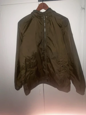 Moncler Olivgrön vindjacka med huva - Olivgrön vindjacka i lätt och glansigt material med dragkedja framtill, två fickor med knappar och justerbar huva. Jackan har resår vid ärmslut och nederkant för en skön passform. Perfekt för blåsiga dagar och enkel att matcha med olika stilar.