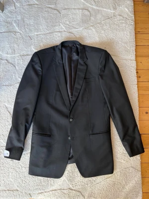 Klassisk svart kostym med två knappar - Säljer en klassisk svart kostym med kavaj och matchande byxor. Kavajen har två knappar, smala slag och tre fickor framtill. Byxorna har raka ben och sidofickor. Perfekt för dig som vill ha en stilren och tidlös look. Du kan även välja att köpa 1 utav dessa för pris vi diskuterar!