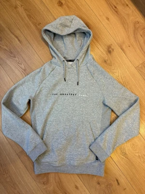 Hoodie ”The Greatest Game” Decathlon  - Snygg gråmelerad hoodie från The Greatest Game med huva och dragsko. Tryck i vitt på bröstet. Tillverkad i mjukt sweatshirtmaterial och har ribbade muddar vid ärmslut och nederkant. Perfekt för en avslappnad och trendig look.