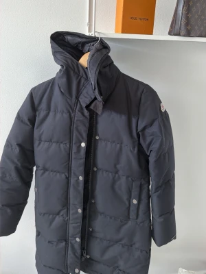 Svart dunjacka från Moncler - Snygg svart dunjacka från Moncler med hög krage och avtagbar huva. Jackan har diskret Moncler-logga på ärmen, dragkedja och tryckknappar framtill samt två fickor med lock. Perfekt för kalla vinterdagar och riktigt stilren look.