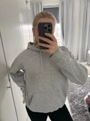 Grå basic hoodie med huva - En stilren grå hoodie med huva och dragsko, perfekt för en avslappnad look. Hoodien har en klassisk känguruficka framtill och ribbade muddar vid ärmslut och nederkant. Tillverkad i mjukt bomullsmaterial för extra komfort.