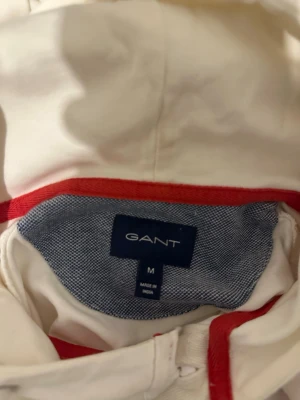 Vit hoodie från GANT med röd detalj - Snygg vit hoodie från GANT med röd panel över bröst och armar där det står 'NEW HAVEN' och 'EAST' i marinblå text. Tröjan har huva och ribbade muddar. Perfekt för dig som gillar sportig och klassisk stil.