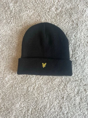 Svart mössa från Lyle & Scott - Säljer en svart ribbstickad mössa från Lyle & Scott med uppvikt kant och den klassiska gula örnloggan broderad framtill. Perfekt accessoar för kyliga dagar och enkel att matcha med olika outfits.