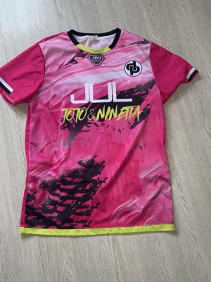Rosa JUL sporttröja Toto & Ninjetta - Snygg rosa sporttröja från JUL med tryck 'Toto & Ninjetta' framtill och nummer 7 samt 'Ninjetta' på ryggen. Tröjan har svarta och vita detaljer, samt neongul kant nertill. Tillverkad i lätt och snabbtorkande material, perfekt för träning eller match.
