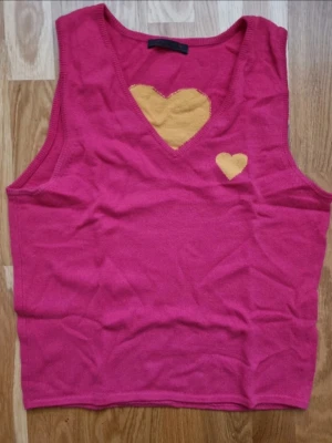 100% kaschmir linnetopp med hjärtan - Söt rosa stickad linnetopp med v-ringning och två gula hjärtan framtill samt ett gult hjärta på ryggen. Ärmlös modell i mjukt material, perfekt för dig som gillar färg och lekfulla detaljer.